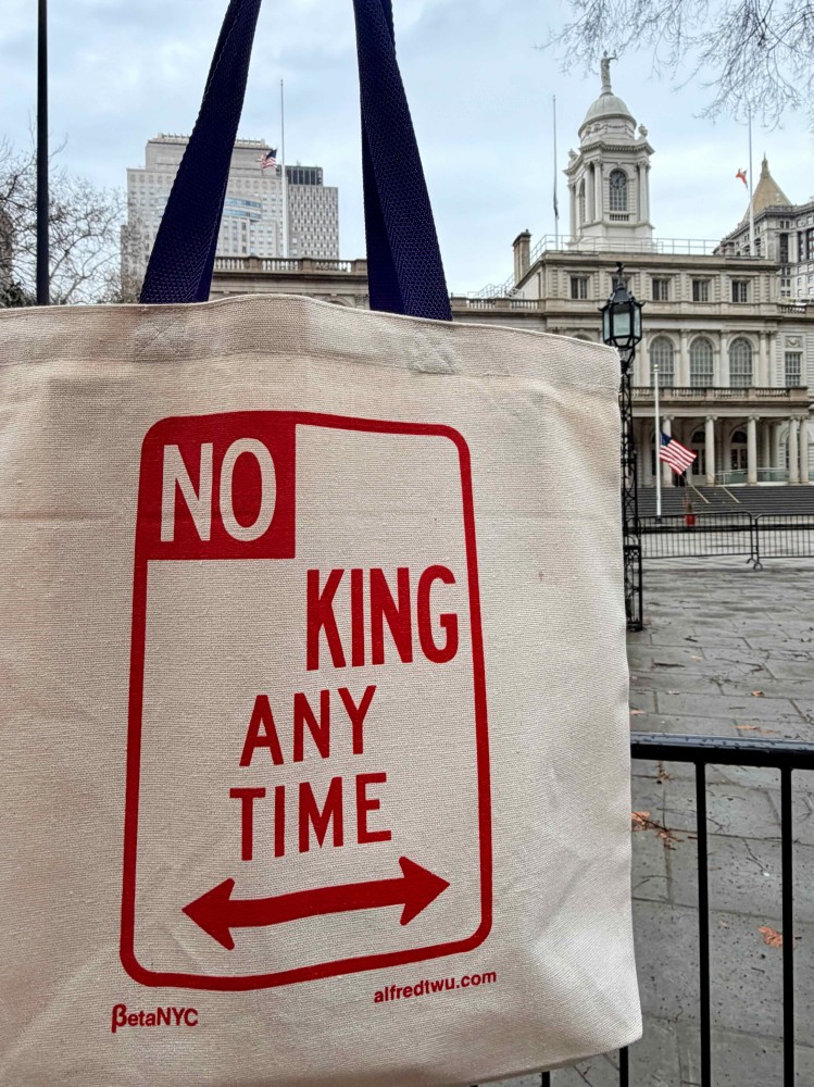 A person holds a beige cotton canvas tote bag outside of NYC City Hall. The bag has navy blue handles and a red logo that reads "NO KING ANY TIME."
