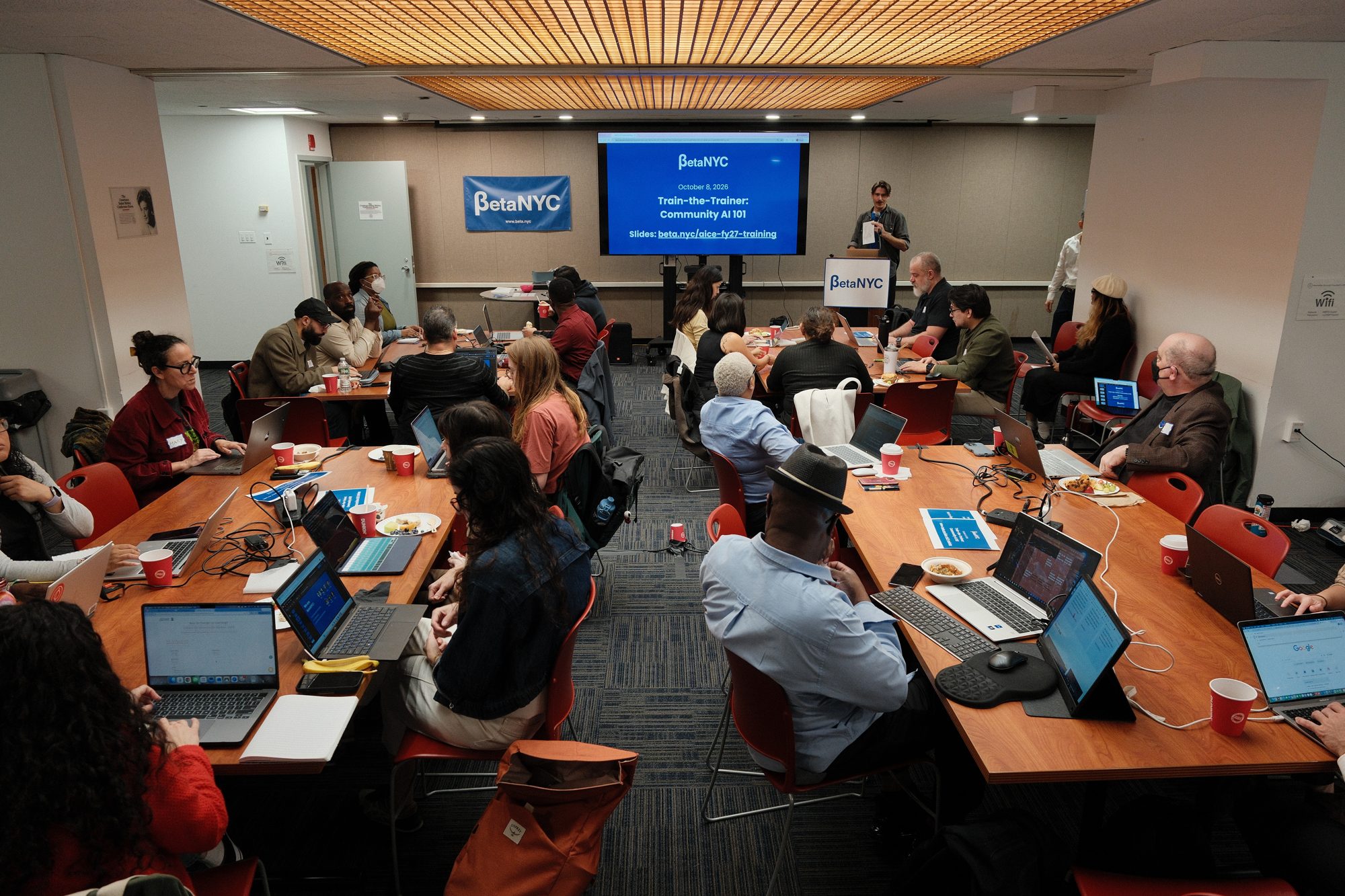 Trainers at tables with laptops in a conference room, with the title slide “Train-the-Trainer: Community AI 101” on the screen at the front.