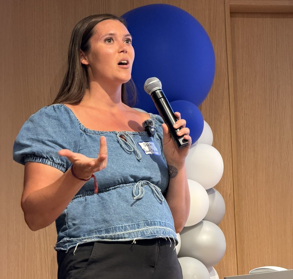 Presenter Ashley Taylor holding a microphone and speaking in front of blue-and-white balloons.