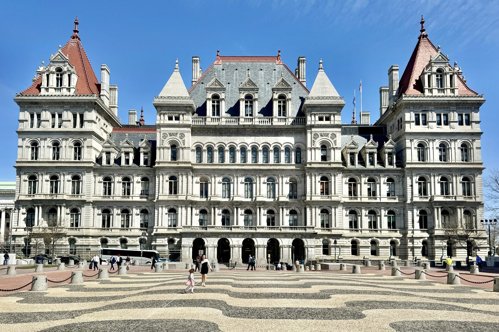 Facade of a stone building with red-tiled towers and arched windows against a blue sky; people walk on a patterned plaza.