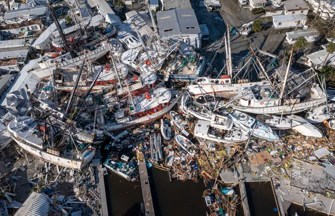 An aerial view showing severe damage to Fort Myers Marina near the bridge to Fort Myers Beach after Hurricane Ian on September 29, 2022. Numerous boats are piled on top of each other amidst debris, surrounded by damaged structures. Photo courtesy of News-Press.