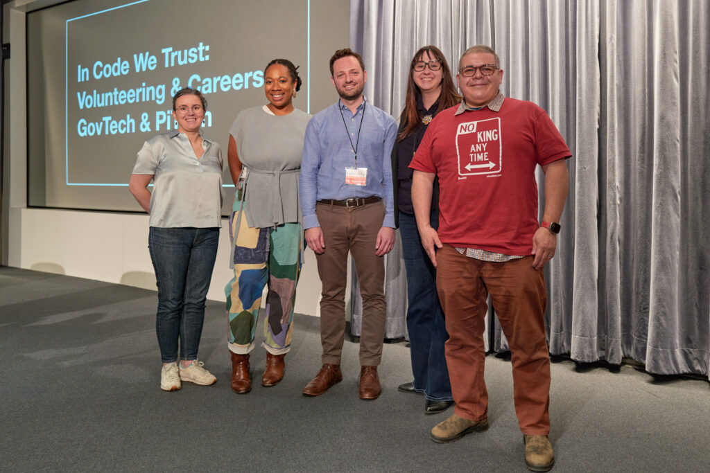 Five panelists stand in a row on a stage, smiling at the camera in front of a large projected slide that reads “In Code We Trust: Volunteering & Careers GovTech & PIT,” with one person wearing a red “No King Any Time” T-shirt.