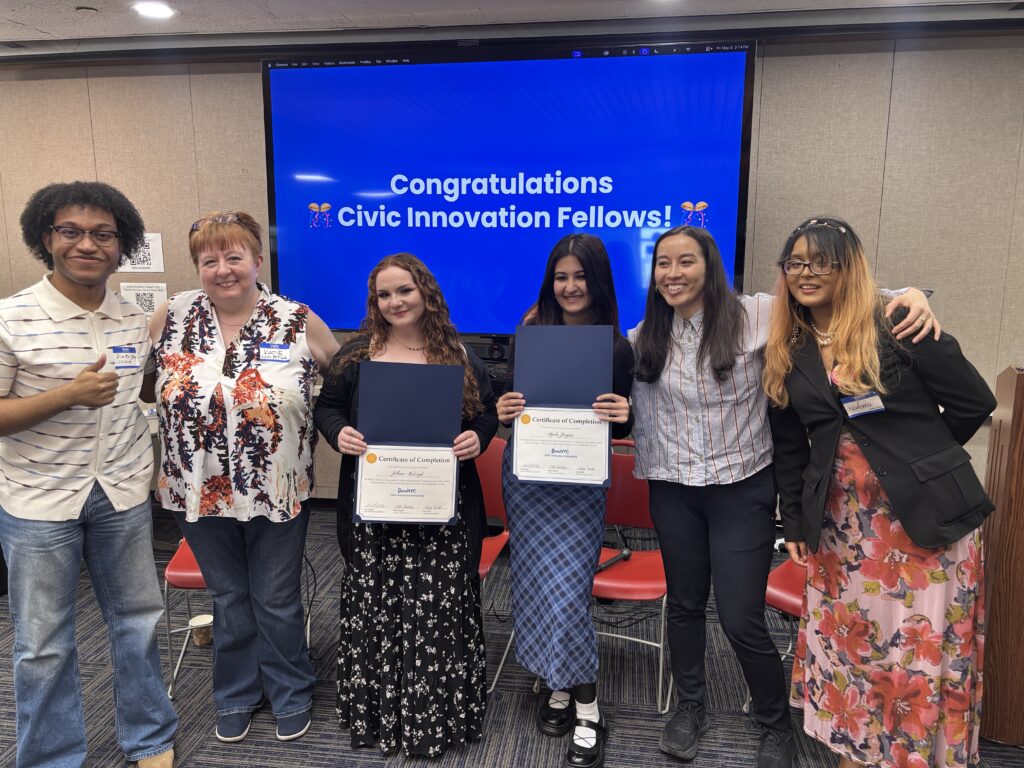Six people stand side by side smiling, with two holding open certificate folders labeled “Certificate of Completion.” A large blue presentation slide behind them reads “Congratulations Civic Innovation Fellows!” indicating a celebratory recognition moment.