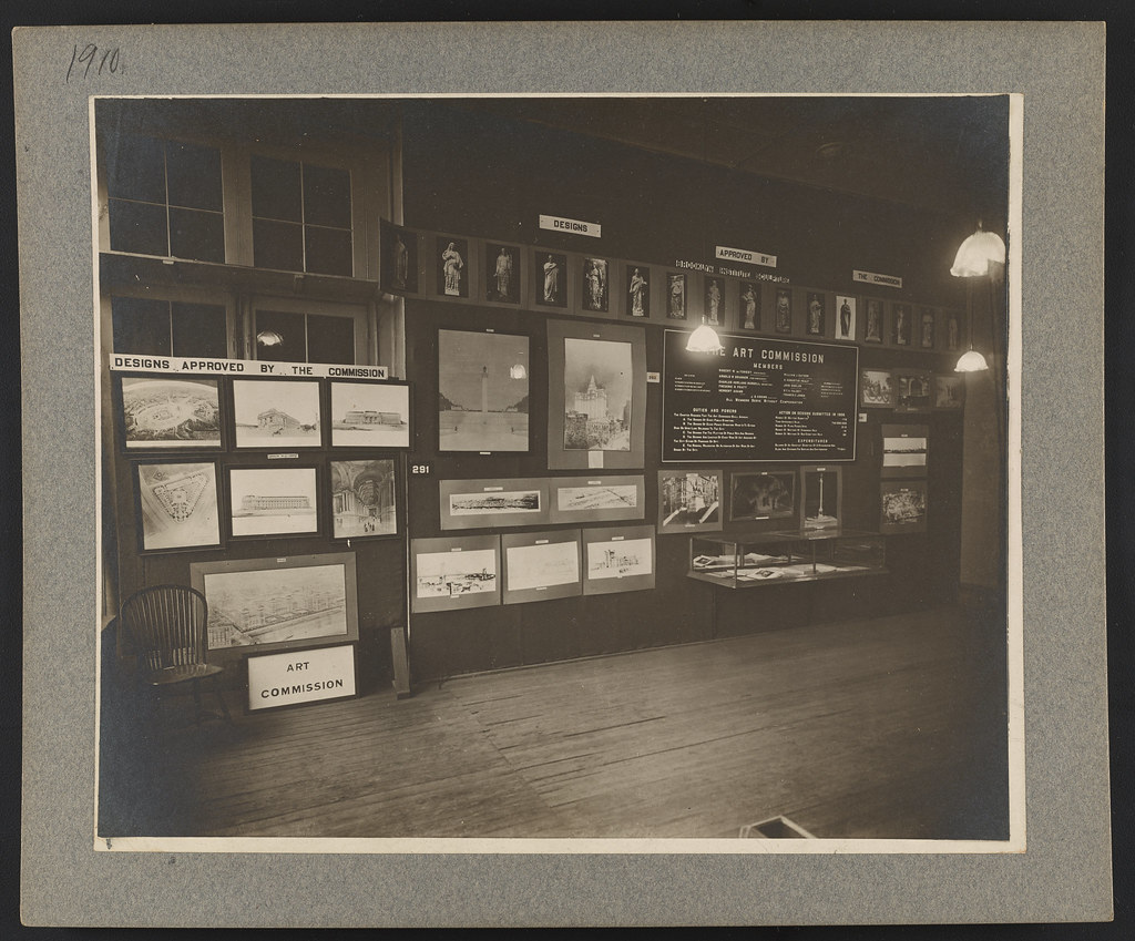 Black-and-white photo of an art commission exhibition with framed design drawings and panels on a wall.
