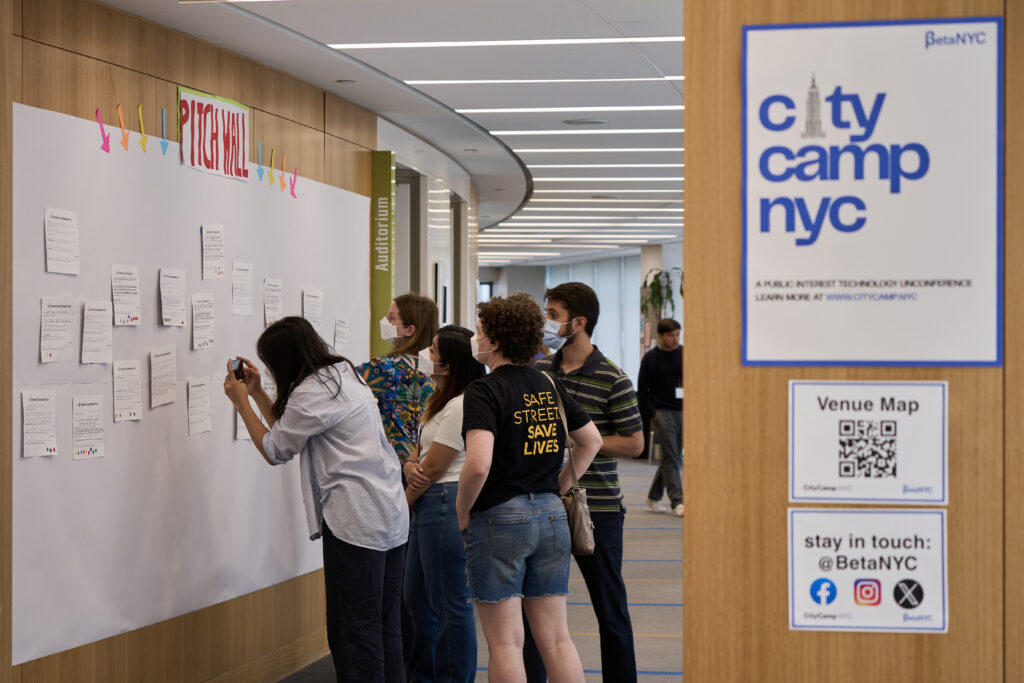 An image of five individuals looking at The Pitch Wall and the sheets of paper with session ideas. One person is taking a photo of a session pitch with their mobile phone.