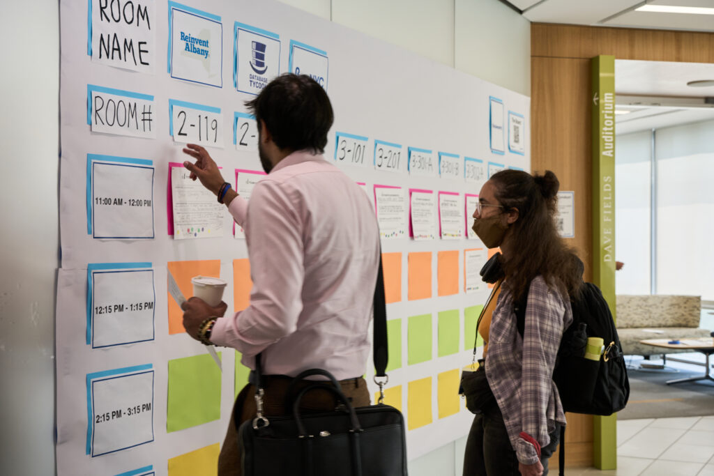 An image of two CityCamp NYC attendees, a man and a woman, studying The Board and looking at room numbers to decide what session to go to