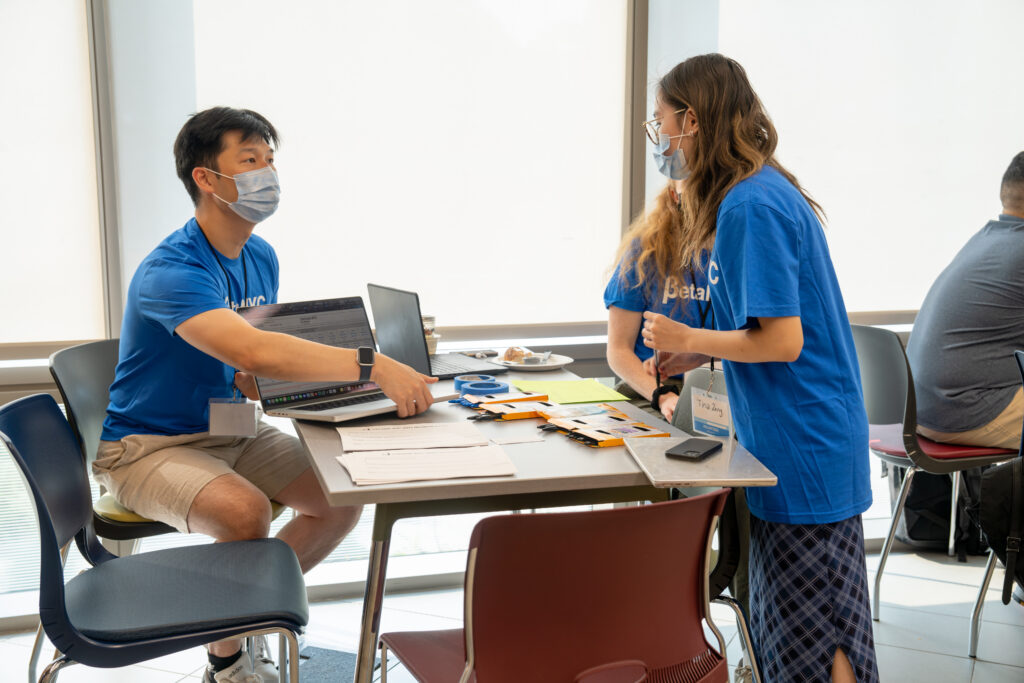 An image of three BetaNYC volunteers wearing bright blue shirts who are conversing together while surrounding a table full of conference supplies