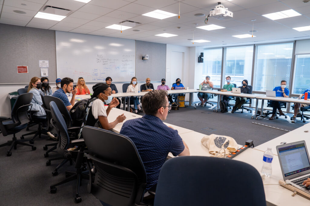 An image of a classroom full of people, seated in a square arrangement where they are all facing each other while discussing a session topic