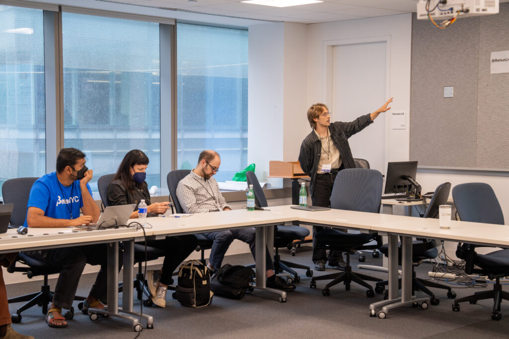 An image of three people studying their laptop and mobile devices, while a fourth man stands at the front of the classroom pointing to a whiteboard.
