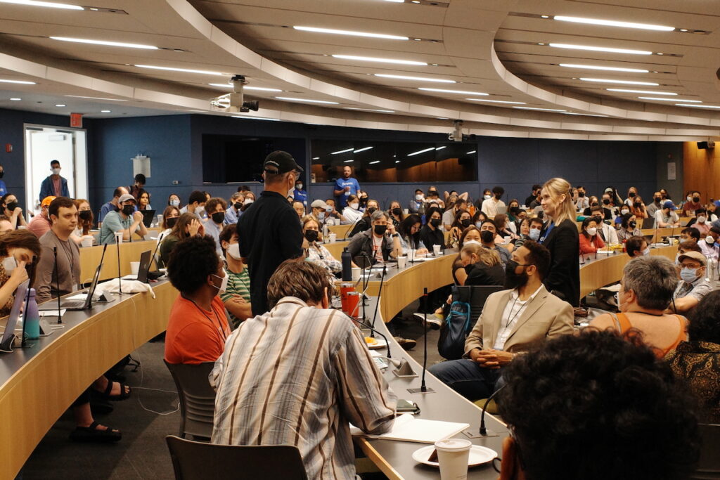 An image of a full auditorium where attendees are listening to one person giving a session pitch