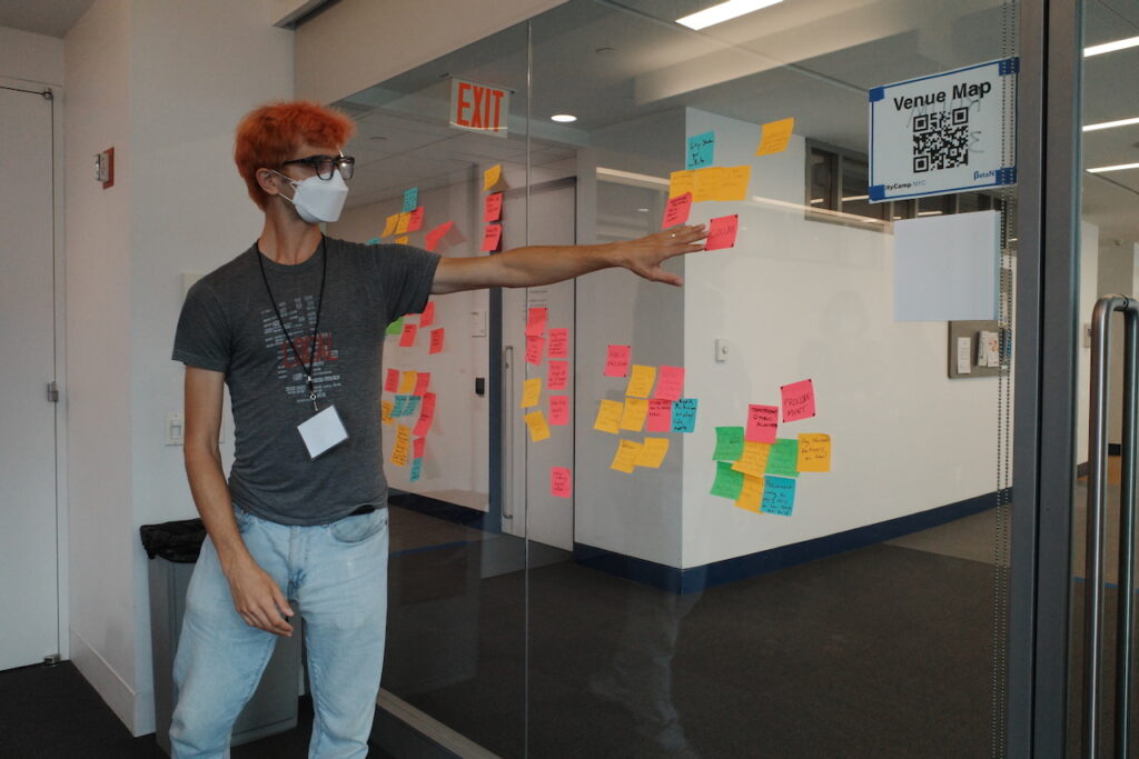 An image of a person wearing kn95 mask with bright pink hair and glasses. The person is pointing to a series of sticky notes on a glass window