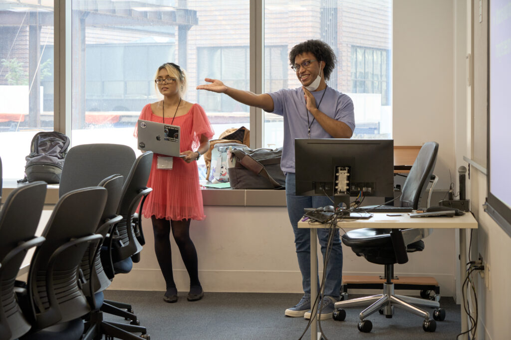 Two presenters stand at the front of a computer classroom; one holds a laptop while the other gestures enthusiastically toward a projected screen, with rows of empty chairs and large windows behind them.
