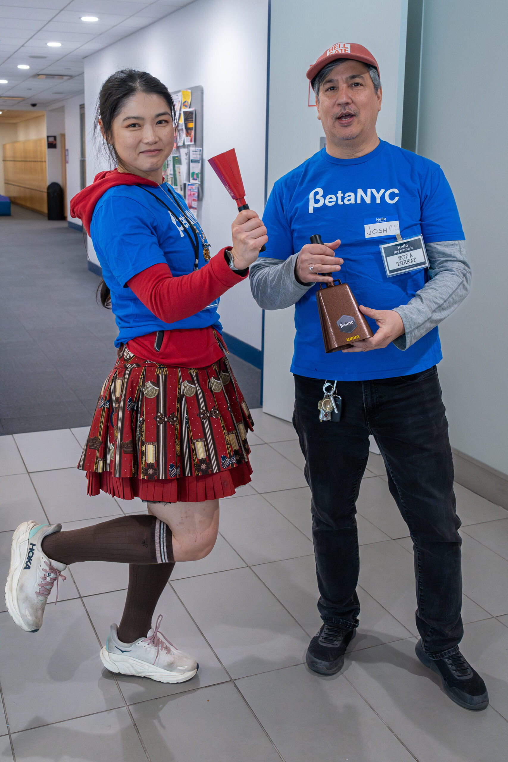Two BetaNYC volunteers pose at the entrance of a building hallway. The woman on the left, holds a red small hand-held bell and poses playfully with one foot kicked up. The man on the right, with a red "HELL GATE" hat, holds a larger bronze-colored bell and wears a blue "BetaNYC" t-shirt and name tag. They are both smiling.