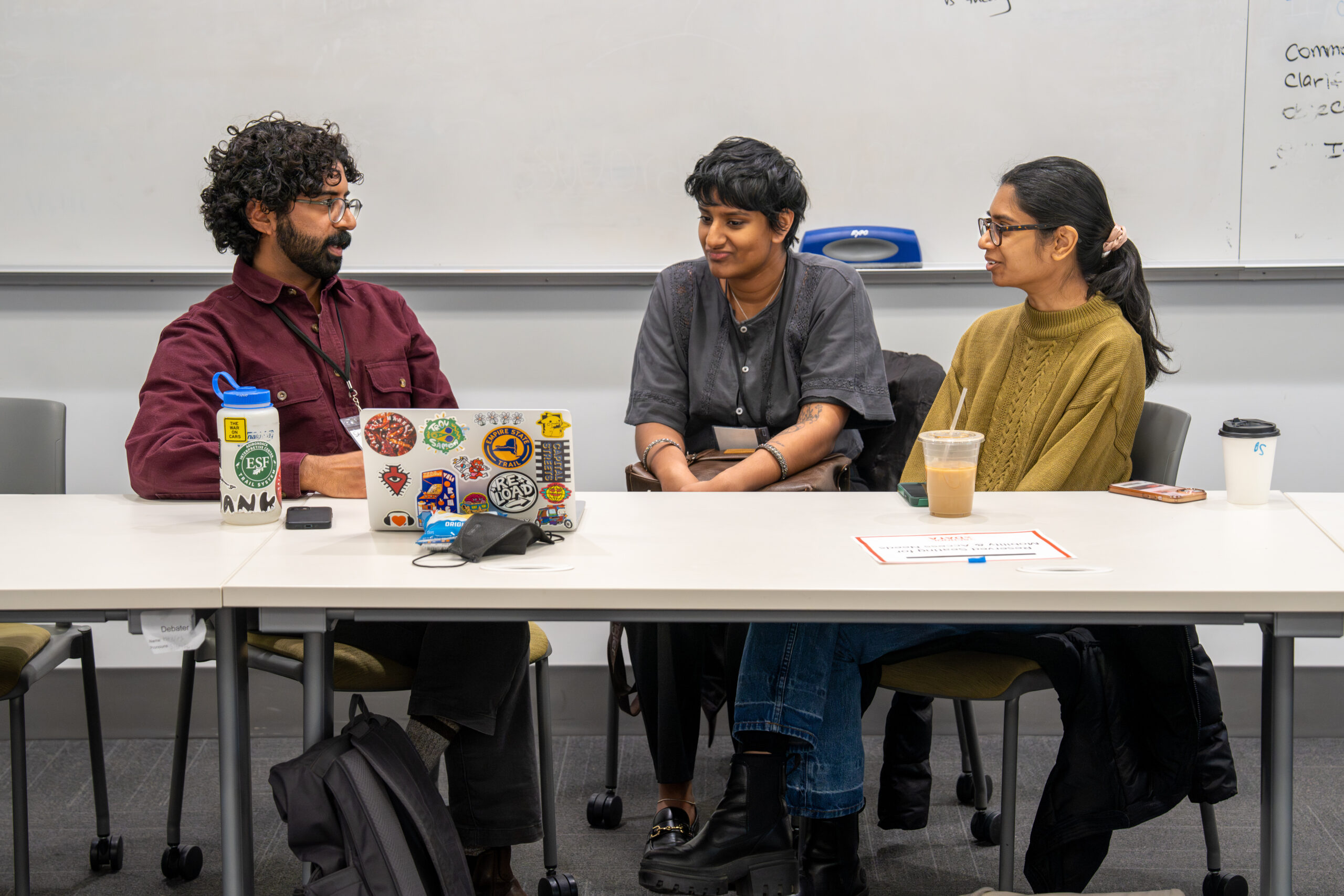Three people are seated closely behind a table, deeply engaged in conversation. The person on the left speaks while the two on the right listen attentively. A sticker-covered laptop sits open in front of them.