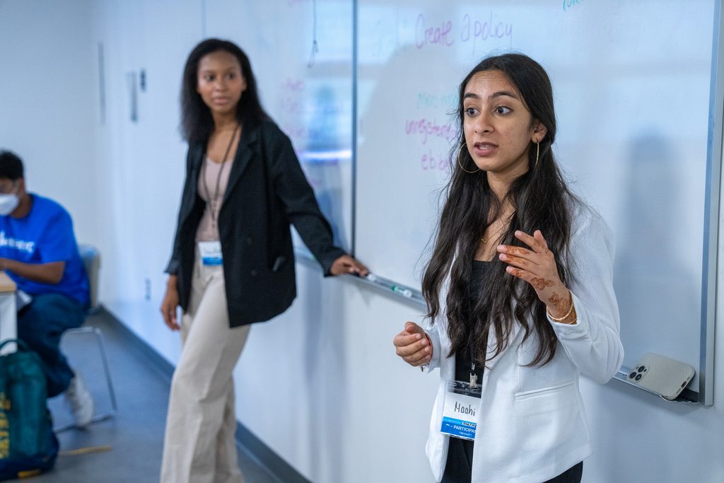 Two presenters lead a session at a whiteboard titled "Create a policy."