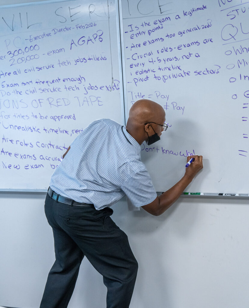 A participant writes on a whiteboard covered in notes about civil service exams and tech jobs.