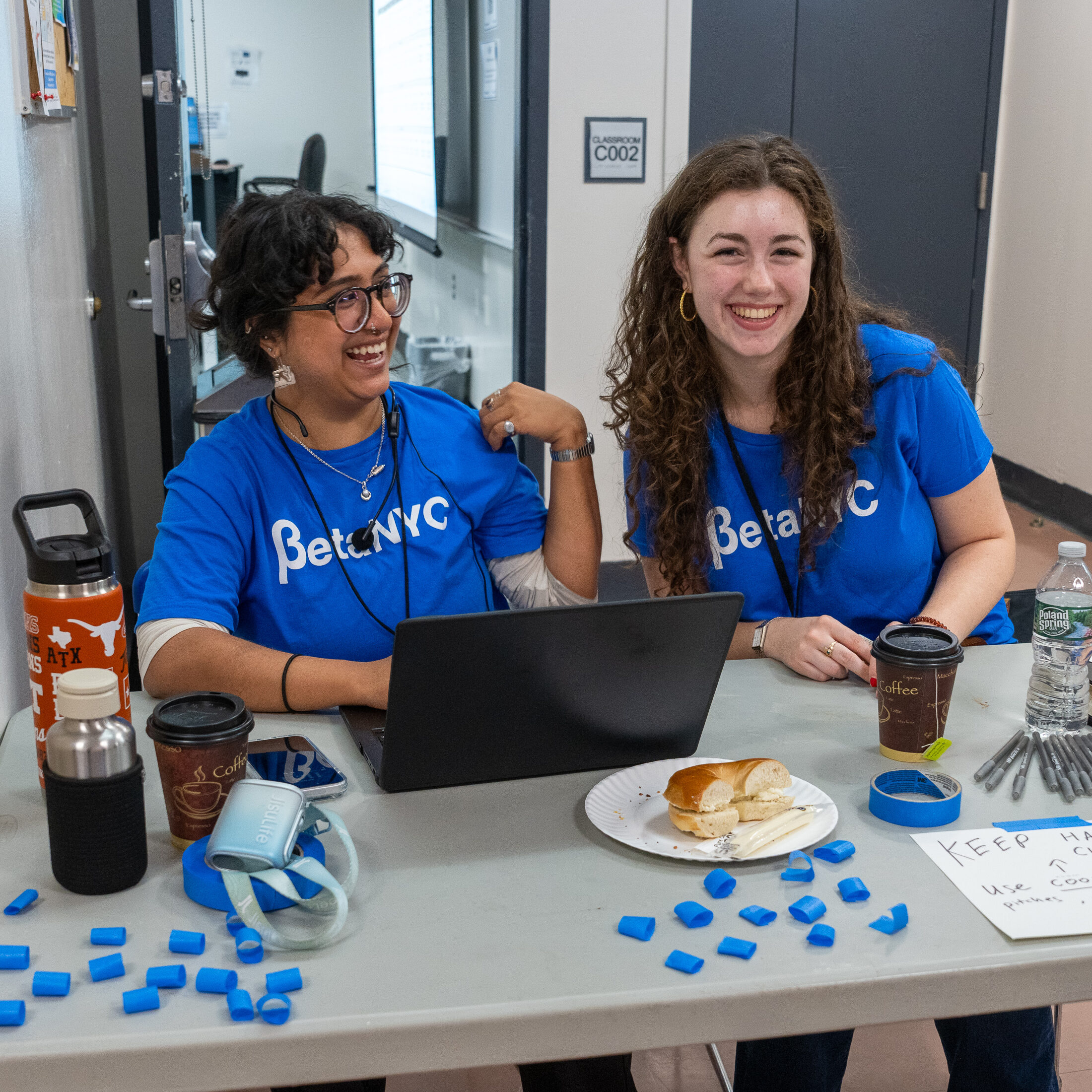 Two smiling volunteers in BetaNYC shirts staff a The Board Building table with a laptop and supplies.