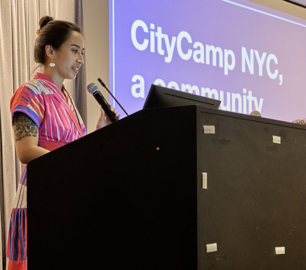An image of Jazzy Smith in a bright, colorful dress, on stage holding a microphone and giving opening remarks.