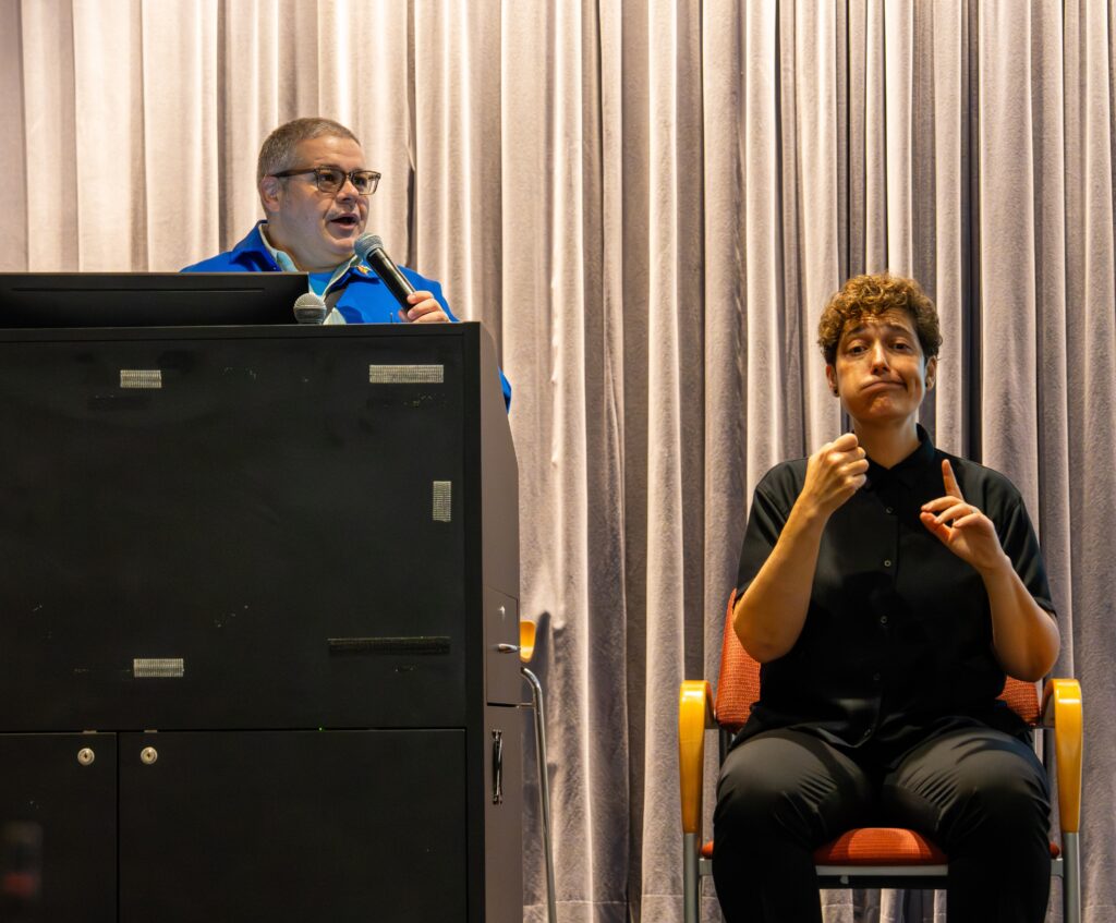 An Image of Noel Hidalgo in a bright blue jacket on stage holding a microphone and giving remarks. An American Sign Language interpreter wearing dark clothes is seated next to him while signing.