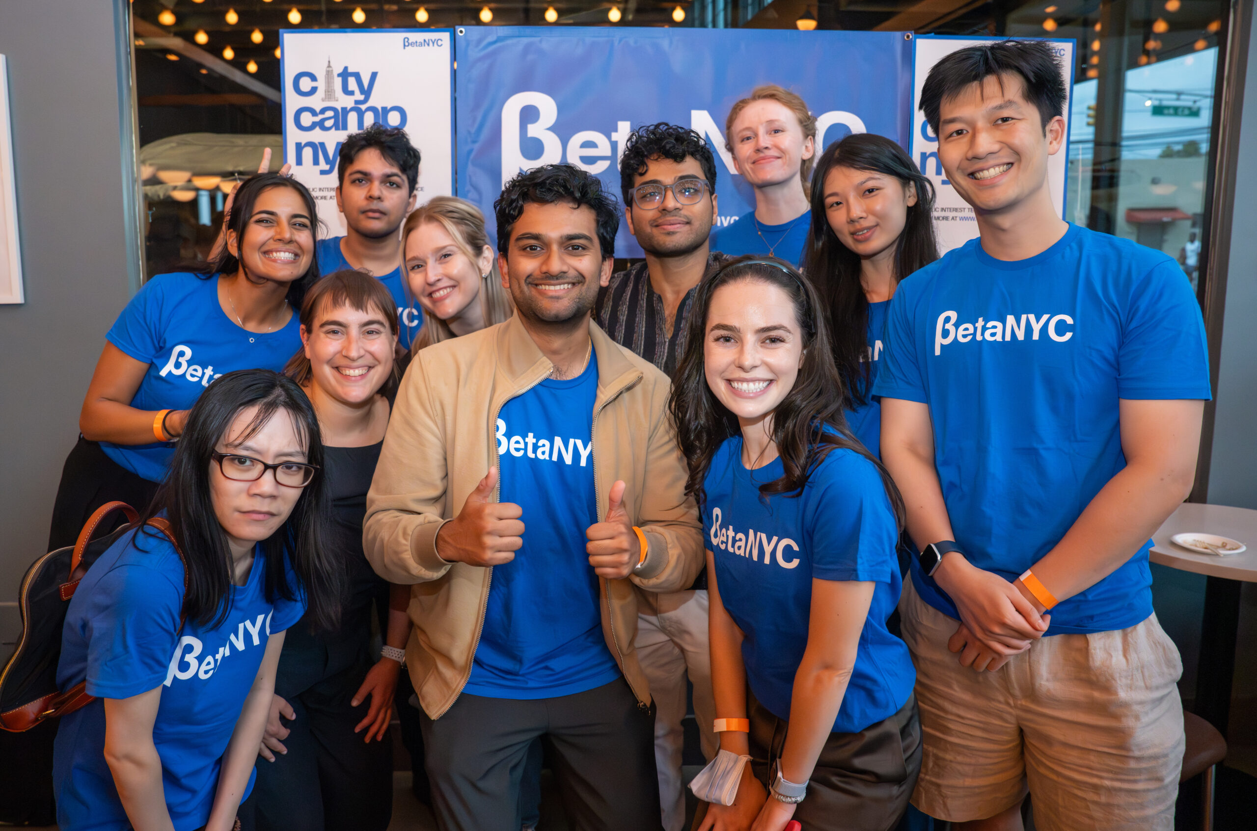 A group of twelve smiling people pose together at a CityCamp NYC event. Most are wearing matching blue “BetaNYC” t-shirts, standing and crouching closely in front of a banner that reads “CityCamp NYC” and “BetaNYC.” Warm string lights and large windows create a welcoming indoor atmosphere.