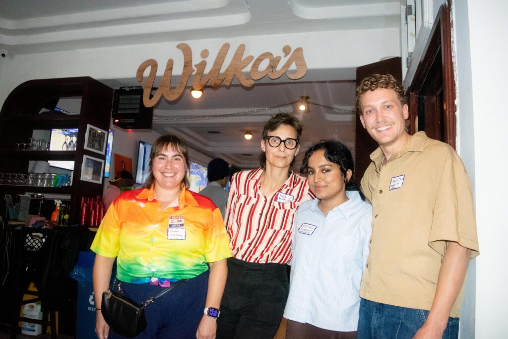 Four adults posing together in a bar setting; rainbow shirt woman, two women in striped shirts, and a smiling man in a beige shirt nearby.