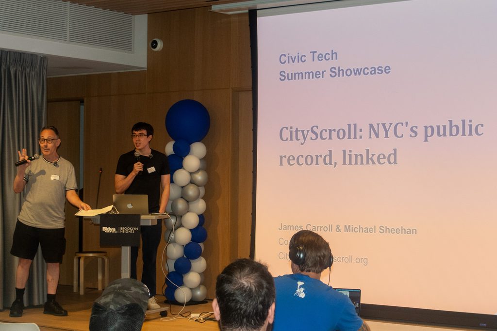 Presenters James Carroll and Michael Sheehan on stage introducing CityScroll, with a slide reading “NYC’s public record, linked.”