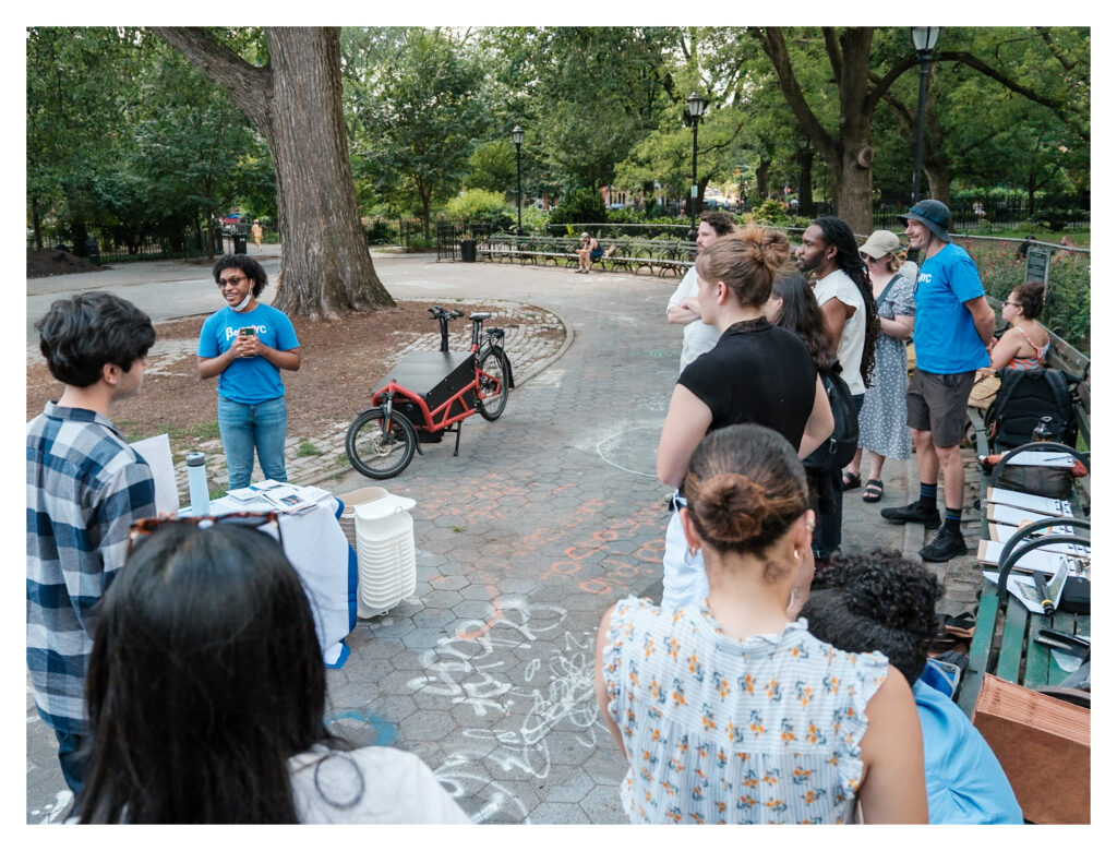 Dimitri at one of BetaNYC's Mapping for Equity events. He is wearing a blue BetaNYC t-shirt and is guiding people on how to map features. They stand in a park alongside benches, trees, and a table. 