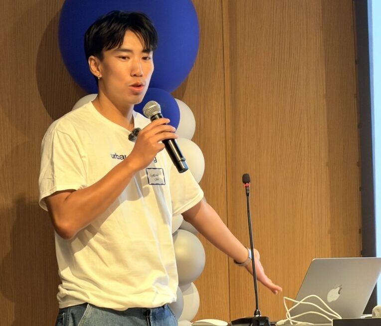 Presenter Gibson Chu speaking in to a microphone next to a laptop and blue-and-white balloons.