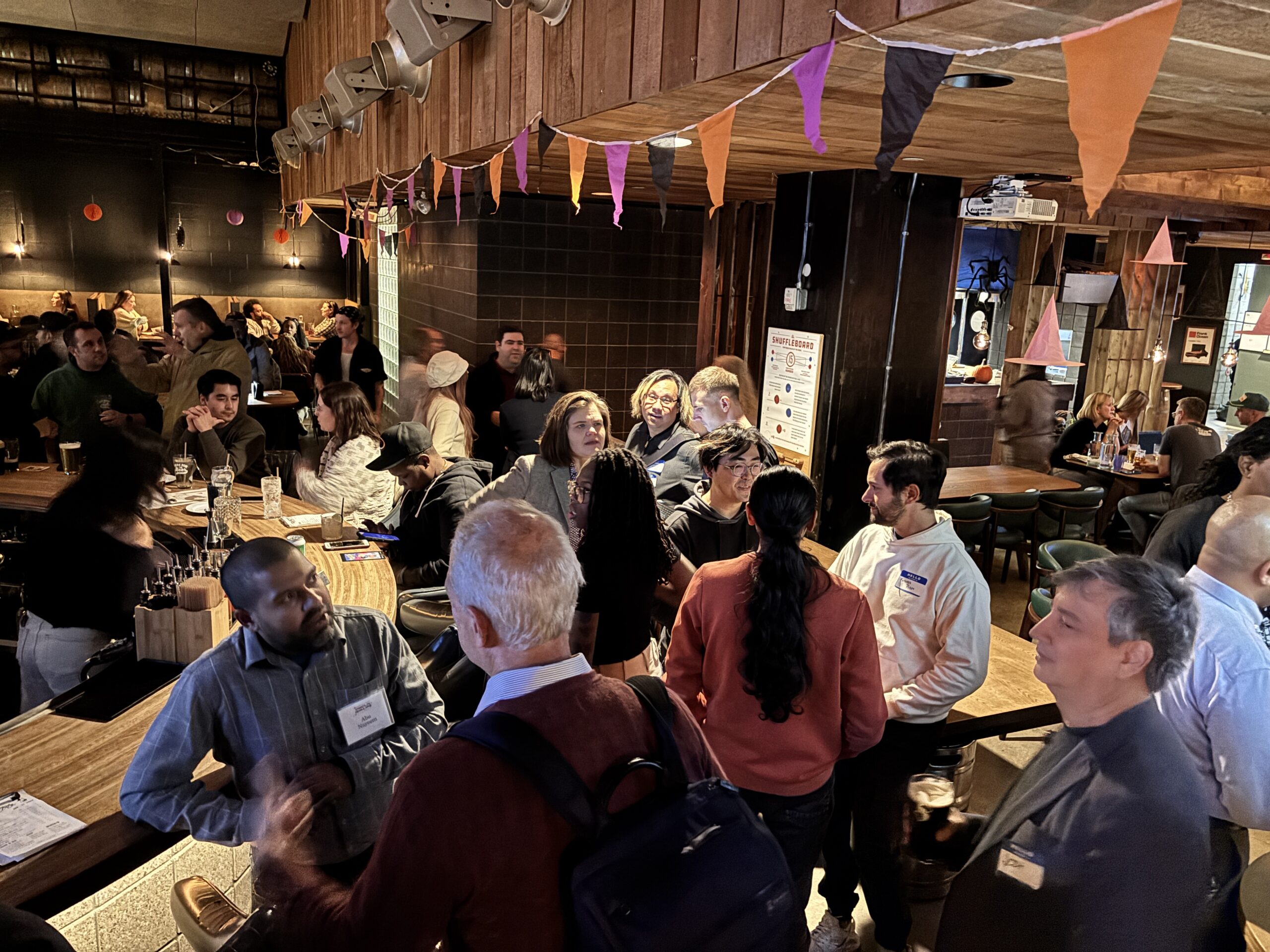 A busy networking event in a wood-paneled bar. People wearing name tags mingle around high tables and booths as festive haloween bunting hangs overhead.