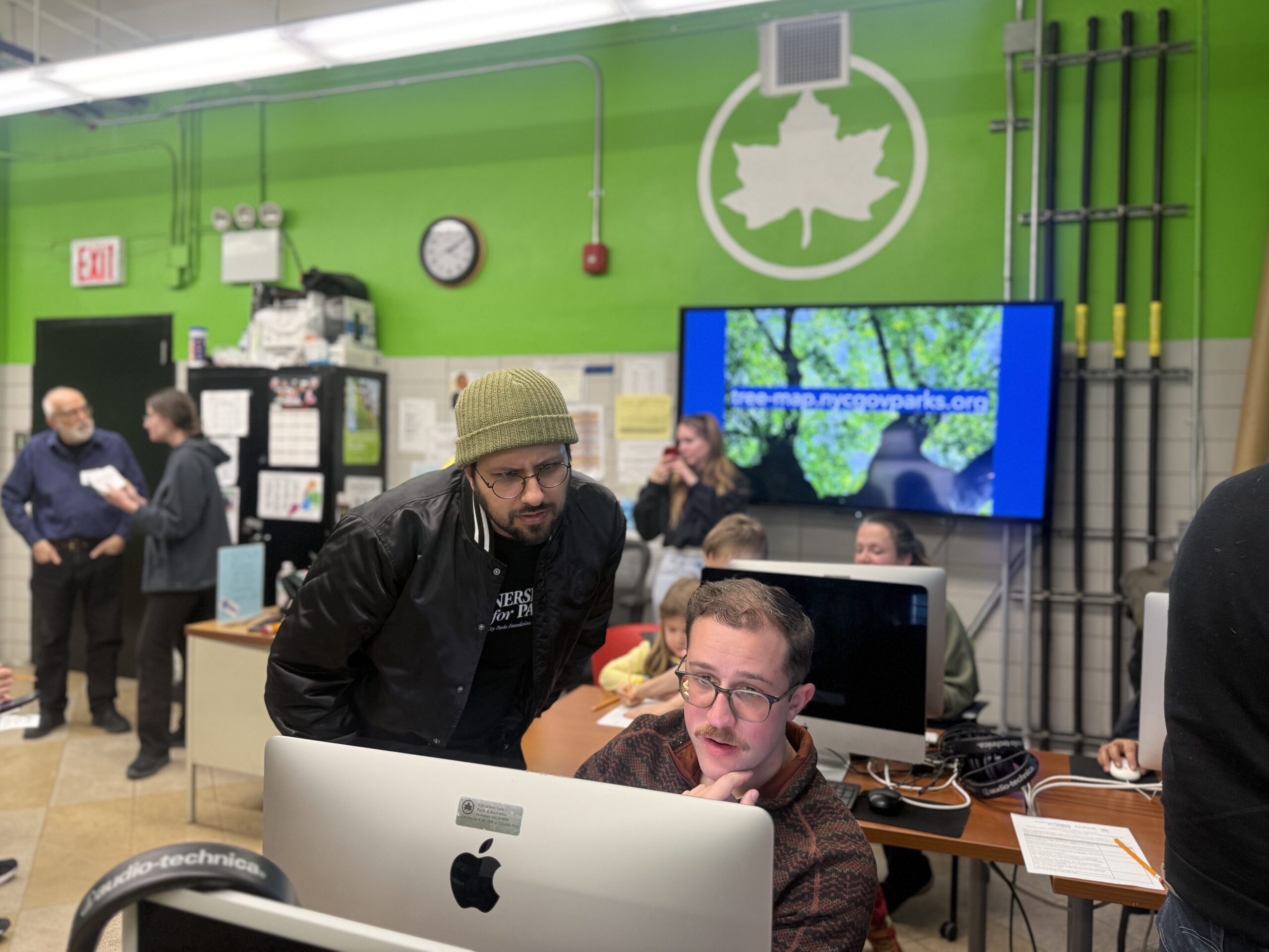 An indoor shot of a brightly lit room with a vibrant lime-green accent wall featuring a large white NYC Parks leaf logo. In the foreground, a man with a mustache and glasses sits at a computer, looking toward the camera. Leaning over him, looking intently at the screen, is another man wearing a black bomber jacket, a green beanie, and glasses.
In the background, other people are working at computers. A television on the wall displays a website for a "tree-map" showing green foliage.
