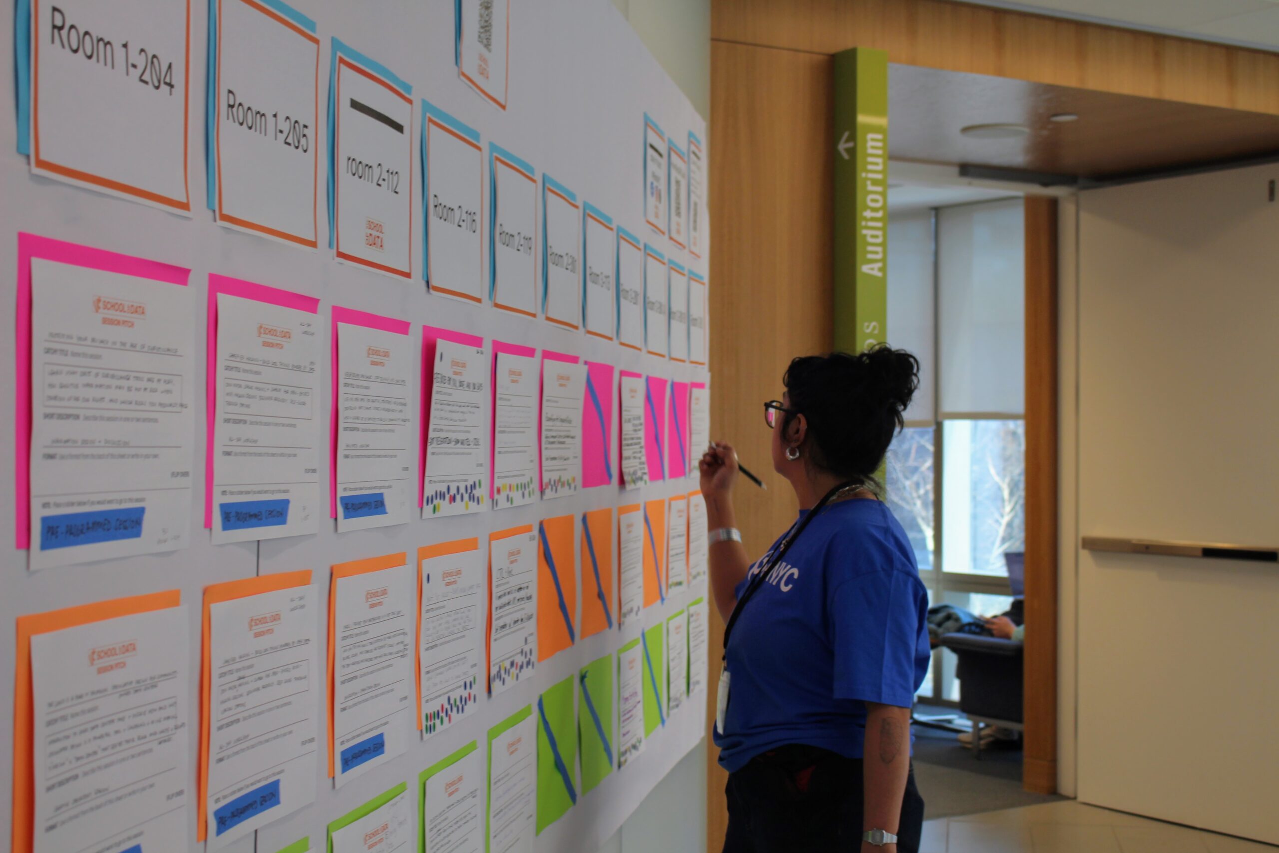 A woman in a blue BetaNYCC t-shirt with stands facing a wall, writing with a black pen on one of many white papers pinned up to the Unconference Session Board