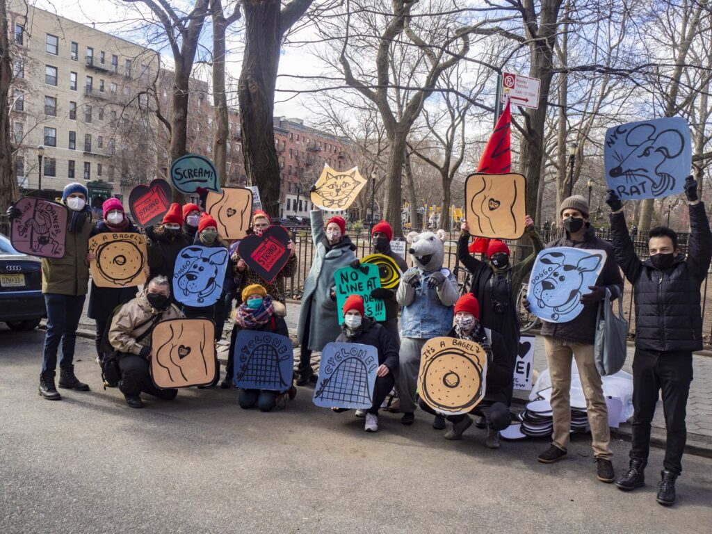 A group of masked people stand together outdoors in a park, holding colorful illustrated signs. The signs feature drawings of bagels, dogs, rats, and abstract designs, with playful slogans. Bare trees and apartment buildings surround them, suggesting winter.