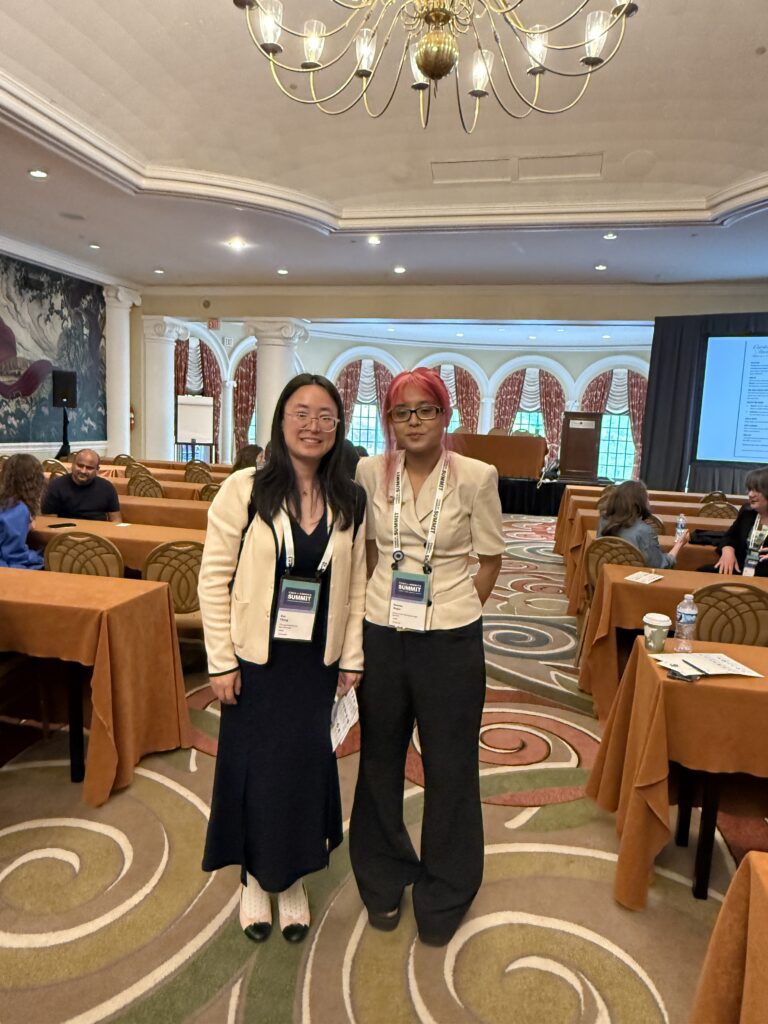 Two woman wearing professional attire and conference badges smile for a photo in a conference room with draped tables, round-backed chairs and presentation screens amongst other attendees.