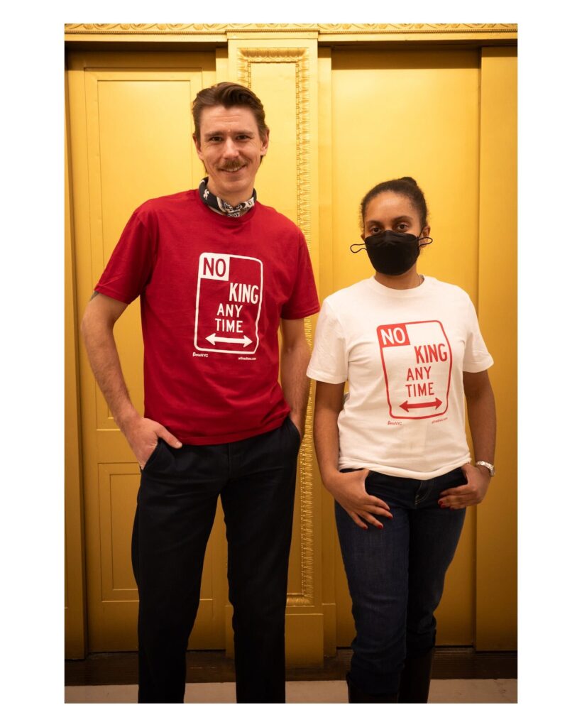 Two people stand side by side in front of gold-colored elevator doors, wearing t-shirts that read "NO KING ANY TIME" in the style of a street parking sign. The person on the left wears a red shirt and a black scarf around their neck, while the person on the right wears a white shirt and a black face mask.