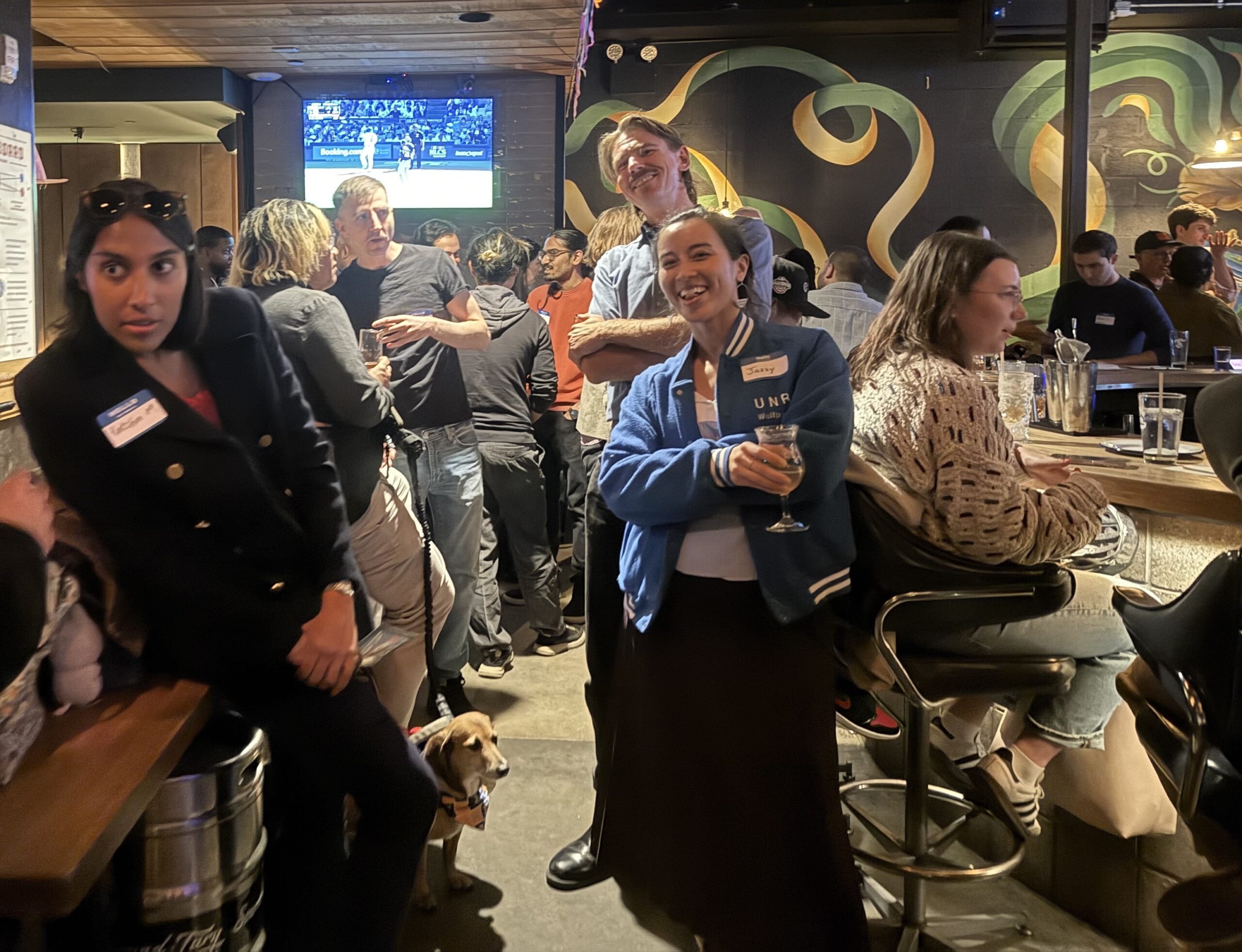 People mingle at a busy bar during a meetup. Two smiling attendees stand at the center with drinks, while others talk nearby; a small dog on a leash rests by the crowd and a TV plays sports in the background.