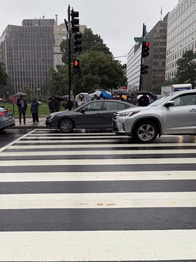 People in business clothing cross a wide zebra-striped crosswalk in Washington, DC, under umbrellas on a rainy day. Traffic and tall buildings fill the background.