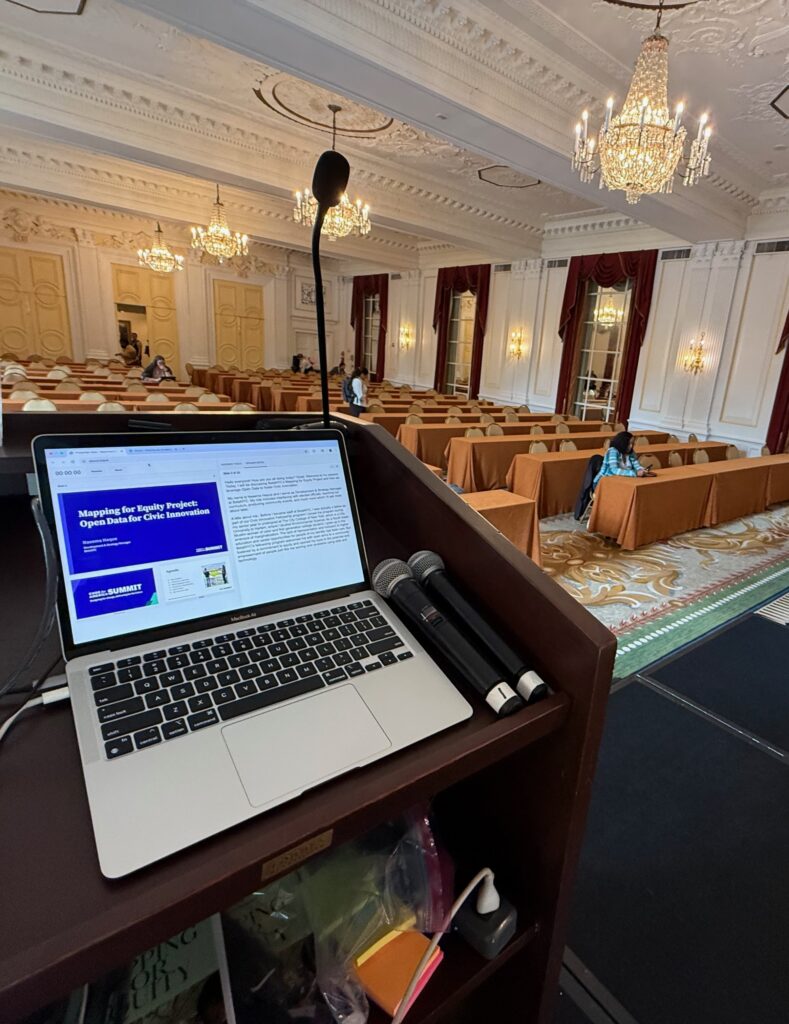 A laptop showing a presentation sits on a podium in an elegant conference room with chandeliers and wood trim.