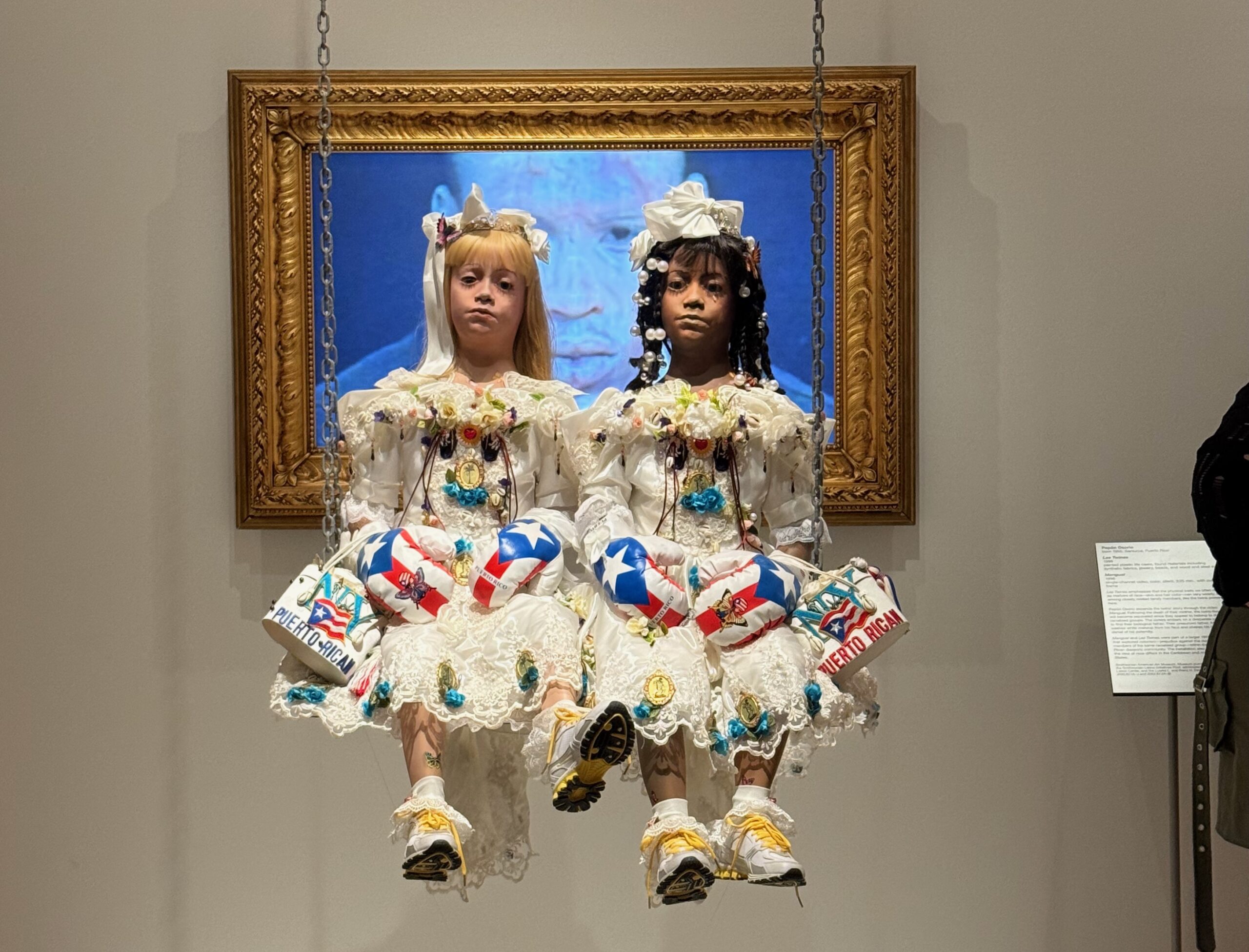 Two hyperrealistic sculptures of young girls with solemn expressions sit side by side on a swing suspended in front of a gold-framed screen. They wear ornate white dresses adorned with colorful embellishments, Puerto Rican flags, and boxing gloves. One girl has light skin and blonde hair; the other has dark skin and black hair with beads. Sneakers peek out from beneath their dresses, blending tradition and contemporary elements.