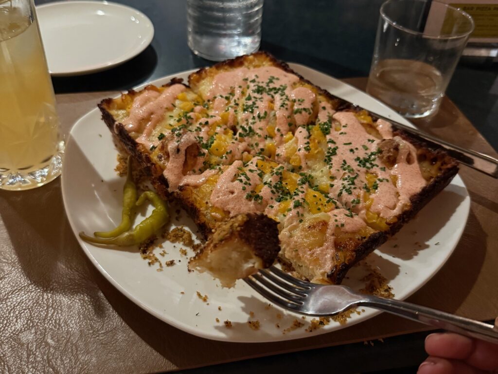  close-up of a rectangular slice of Detroit-style pizza topped with creamy sauce and garnishes, served on a white plate with pickled peppers and a drink in the background.