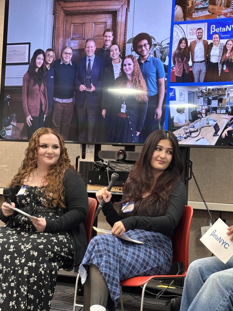 Two panelists sit with microphones during a BetaNYC event. Behind them, a large screen displays a collage of group photos from BetaNYC programs and events, including people posing with an award and participating in meetings.