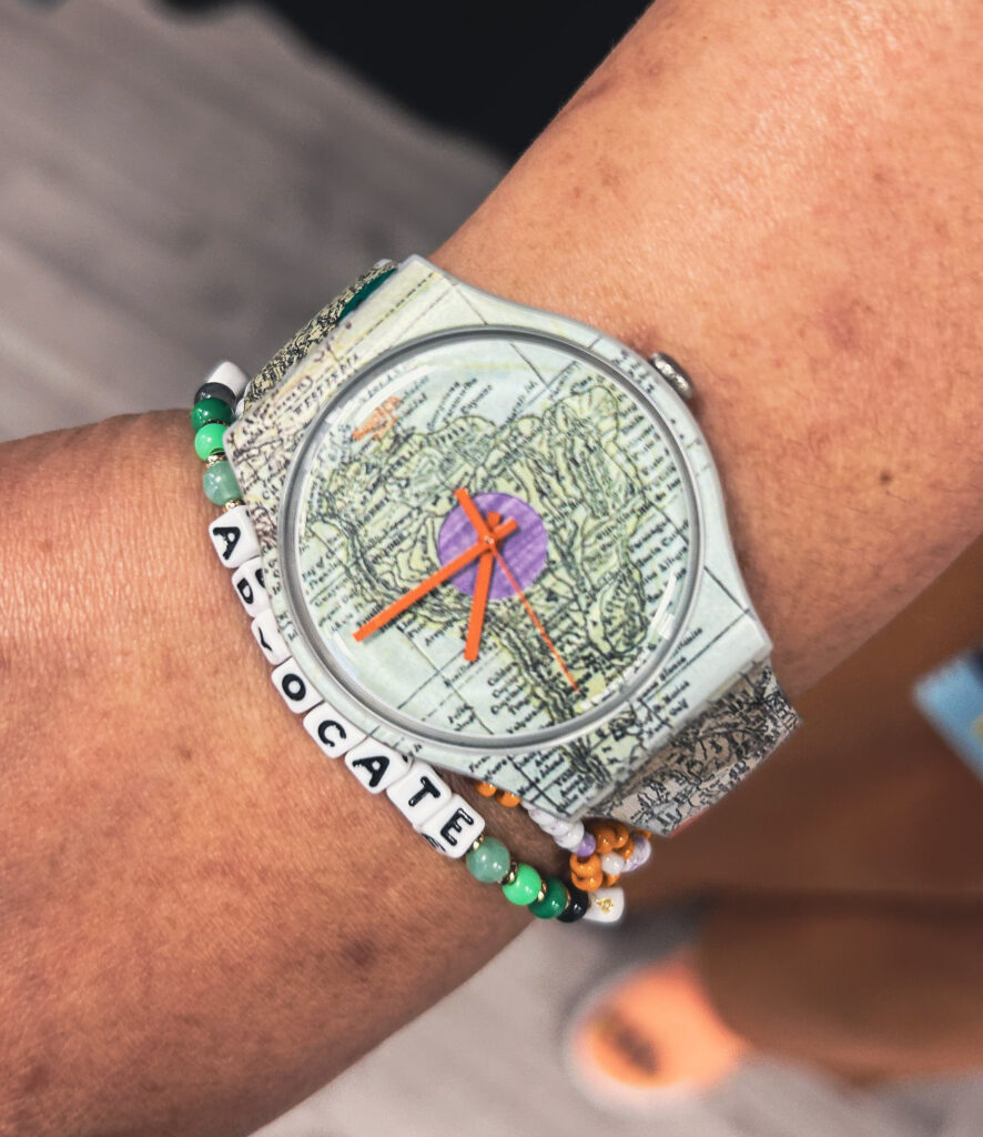 A close-up of Silvia's wristwatch with a vintage map design on the face, worn with colorful beaded bracelets, including one that spells “ADVOCATE.”
