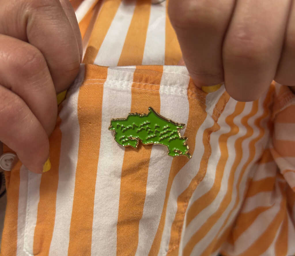 A close-up of a green enamel pin in the shape of Costa Rica, fastened to Silvia's pocket on her orange-and-white striped shirt.