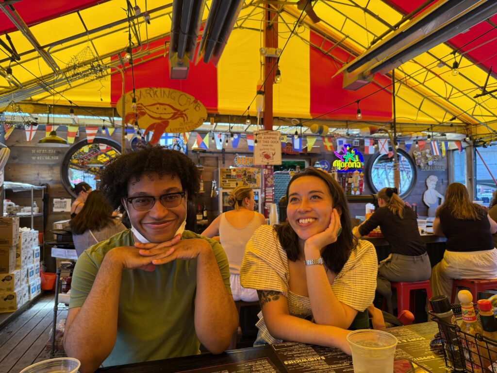 Dimitri and Jazzy sit at a table and pose with their hands. On the table are menus and condiments.