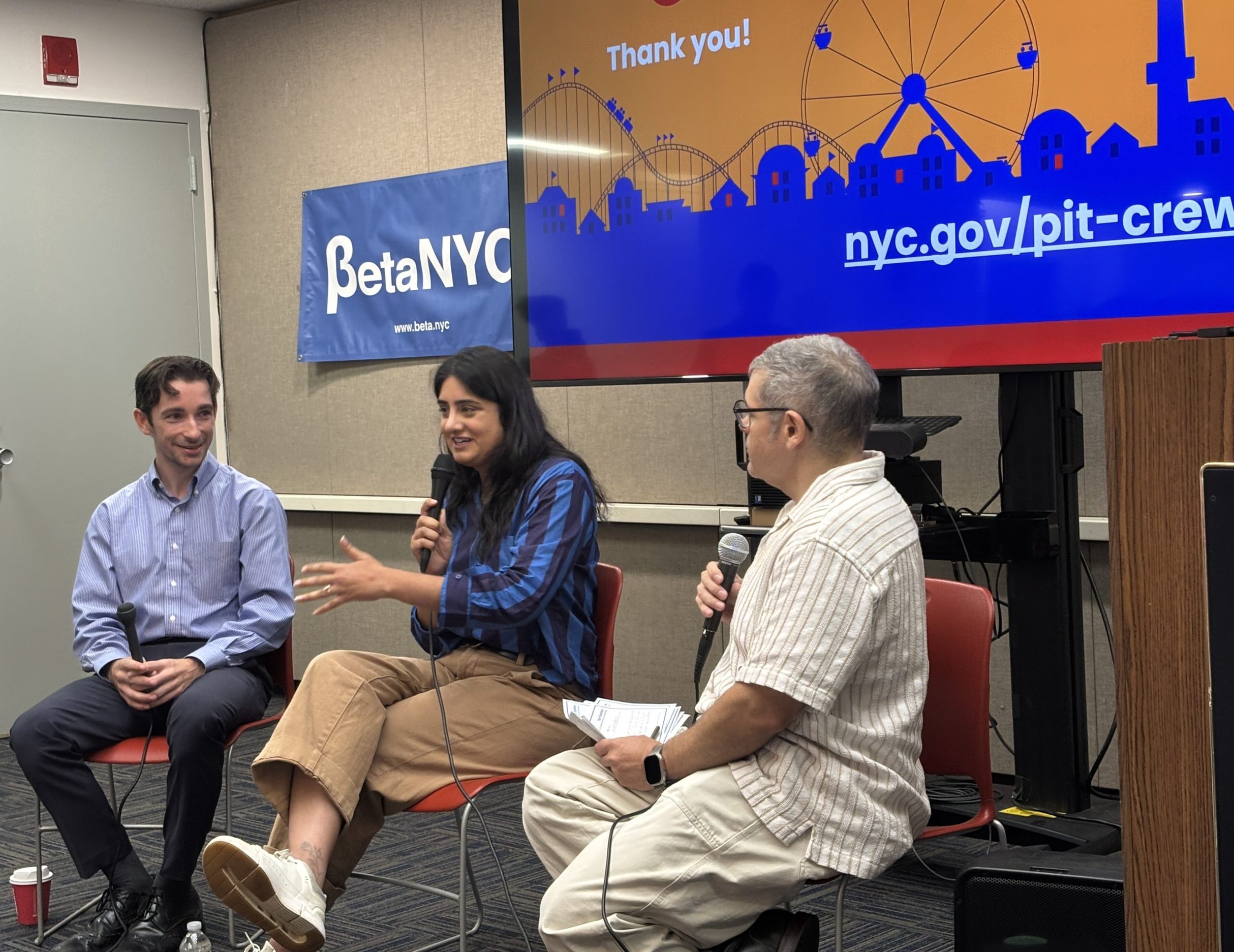 Three panelists seated in chairs, each holding a microphone, indoors with a Betanyc banner in the background and a large orange-blue screen.