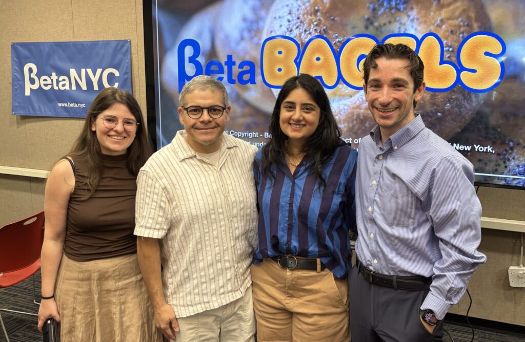 Four people smile for a group photo in a conference room with a large screen showing 'Beta Bakes' and a BetaNYC banner nearby.