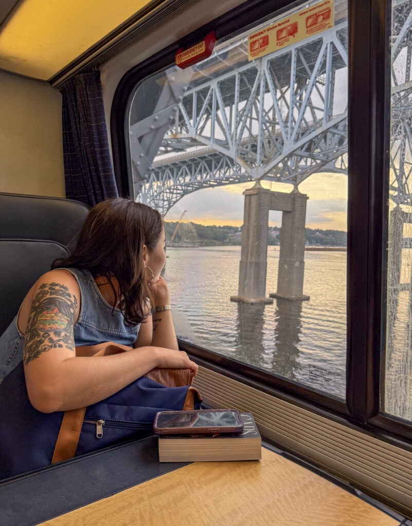 Jazzy with a tattooed arm gazes out the window of a train at a steel bridge over a river, with a phone and book resting on the table.