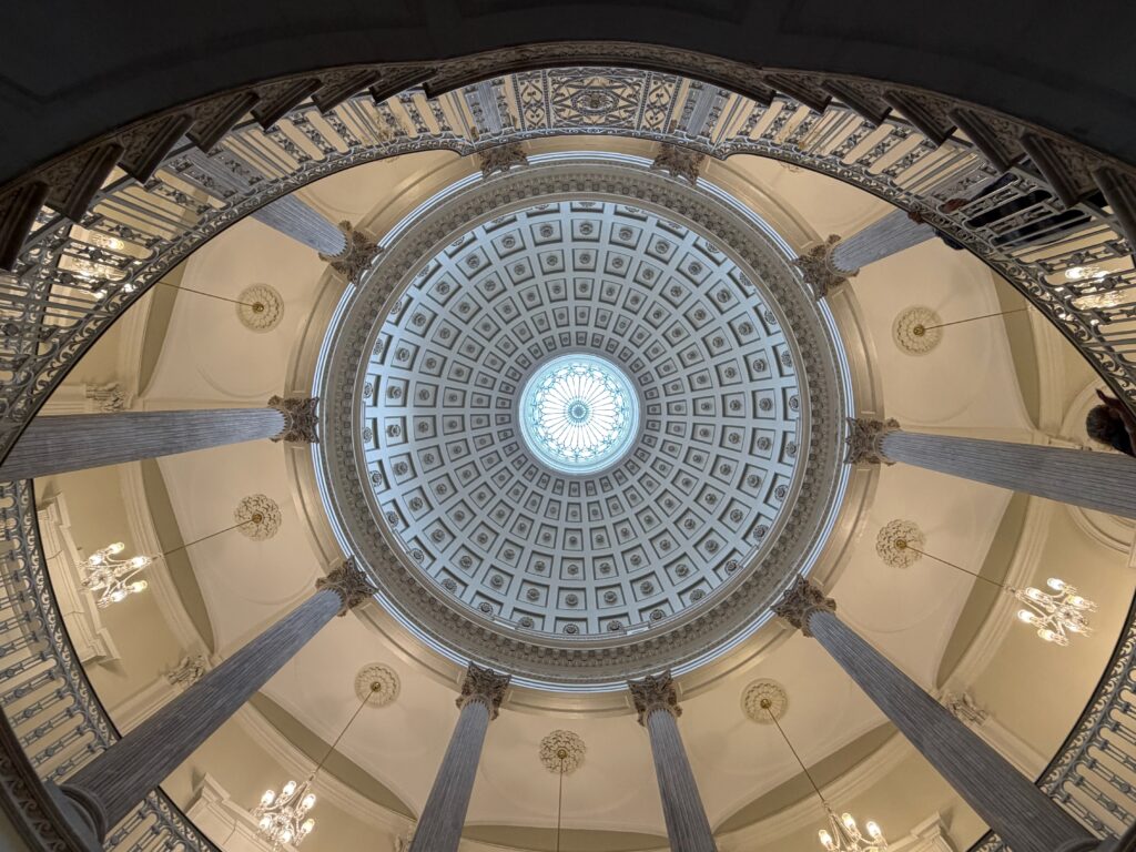 NYC's City Hall Dome — An upward wide-angle shot of a grand neoclassical rotunda interior. Ornate coffered dome with a central skylight, ringed by tall marble columns, decorative moldings, and gilded details. Warm cream tones contrast with the soft blue light filtering through the dome's oculus.