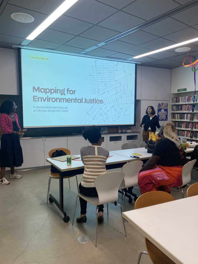 A classroom setting during Climate Week NYC 2024, featuring a presentation titled “Mapping for Environmental Justice: A Geospatial Learning Lab.” Two presenters stand at the front of the room next to a large screen displaying the title slide with a map in the background. Several participants are seated at tables, attentively engaging with the session. The room is well-lit, with bookshelves and colorful decorations visible in the background.