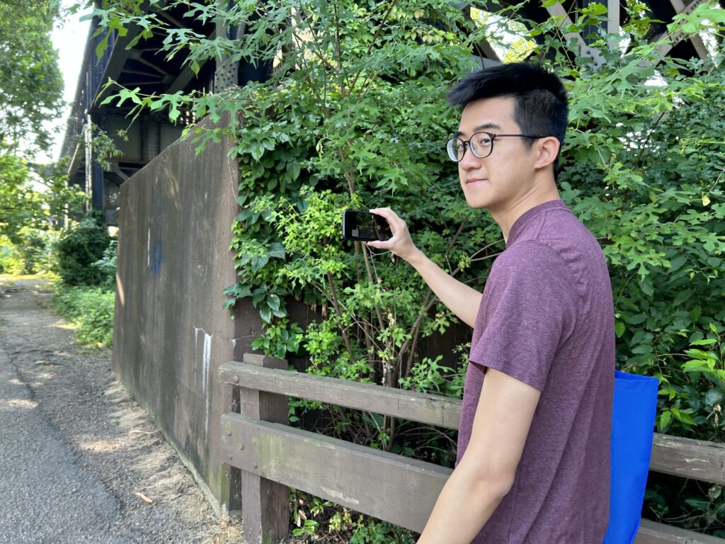 Zhi is in a maroon shirt and glasses stands outdoors beside dense greenery, holding up a smartphone to take a Mapillary footage near an overpass.