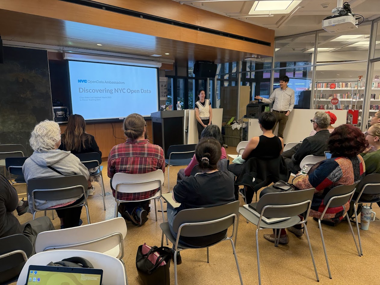 A public library workshop in session—about 15 adults sit in rows facing a screen that reads “NYC Open Data Ambassadors: Discovering NYC Open Data.” A presenter gestures while another stands by the podium; laptops and bags are visible in the foreground. Photo credit: Jane Swanson / Cornell Tech